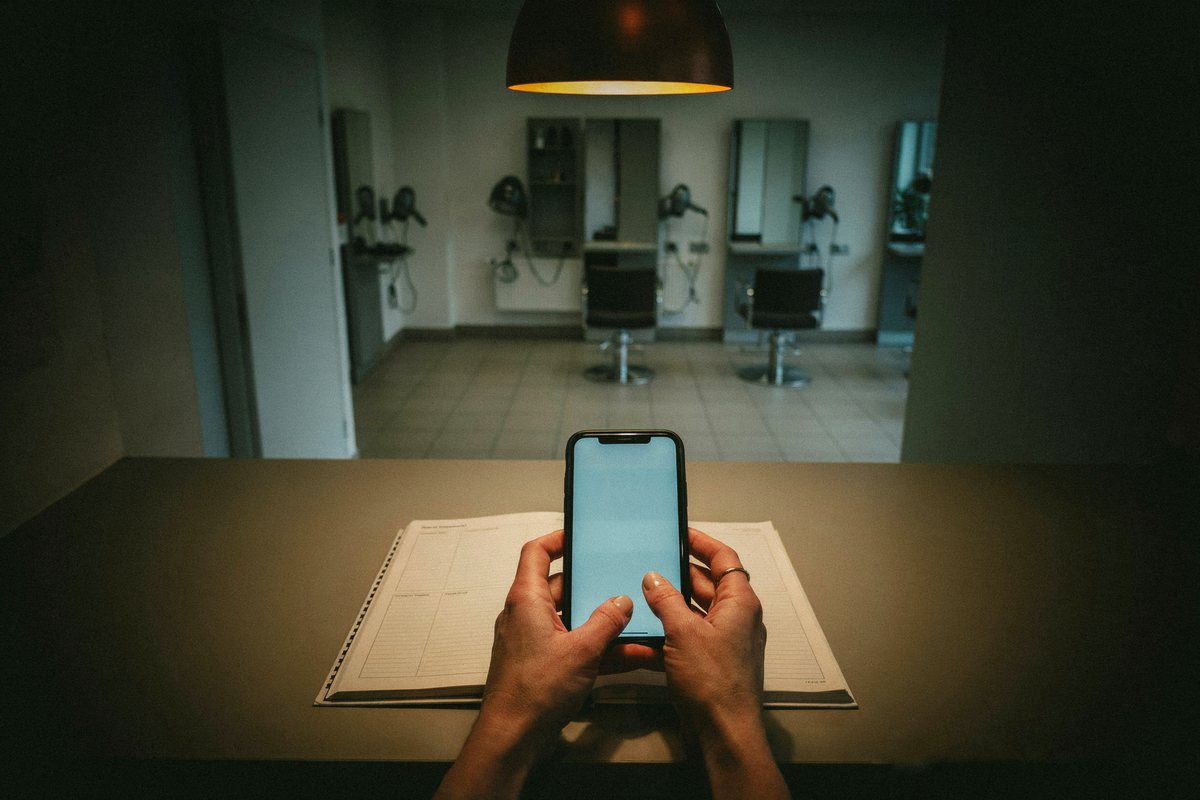 Hände einer Friseurin mit Smartphone über dem Terminbuch im leeren Salon am frühen Morgen