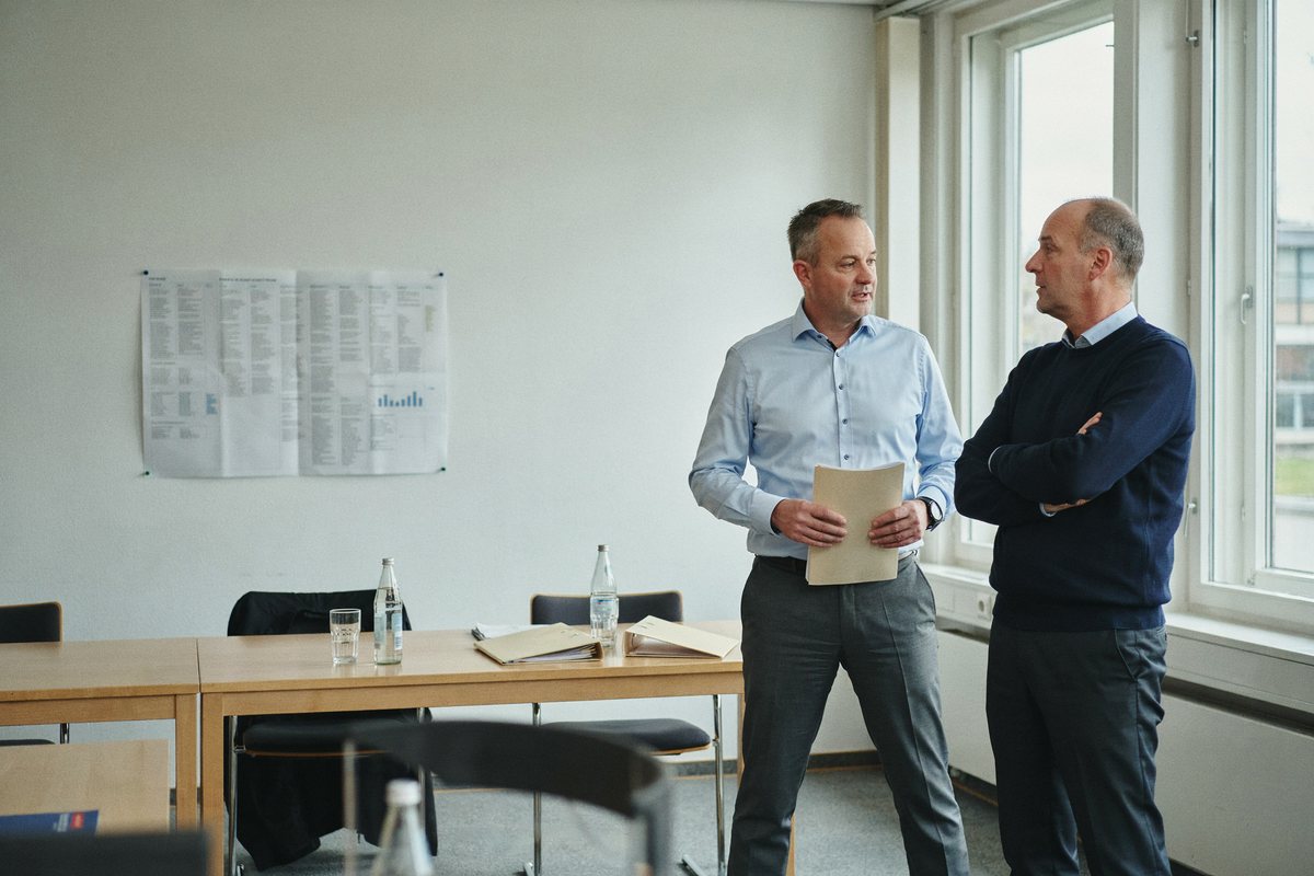 Zwei Führungskräfte im Gespräch am Fenster eines Besprechungsraums, an der Wand ein Strategiepapier