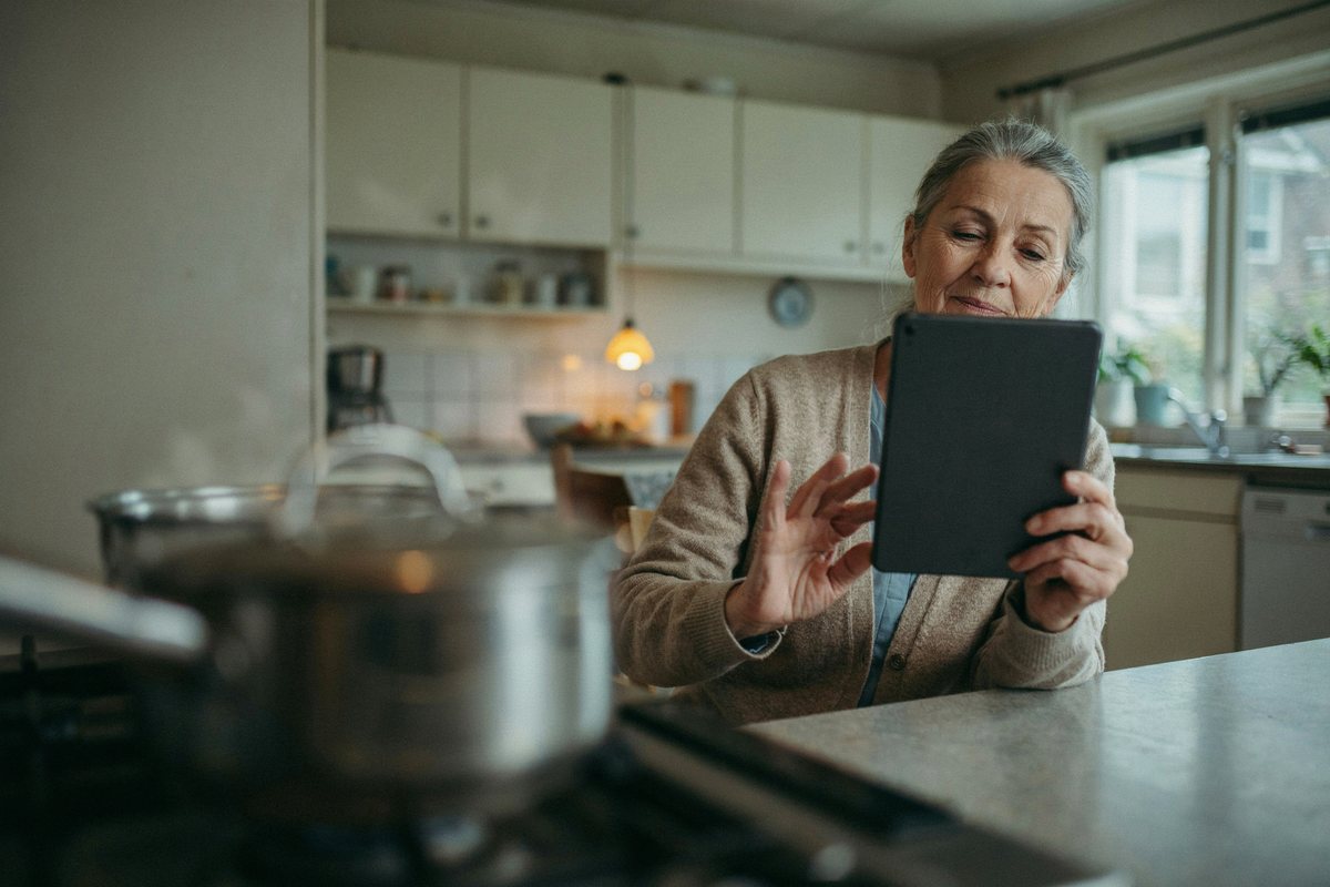 Ältere Frau am Küchentisch spricht mit einem Tablet, im Vordergrund ein Topf auf dem Herd